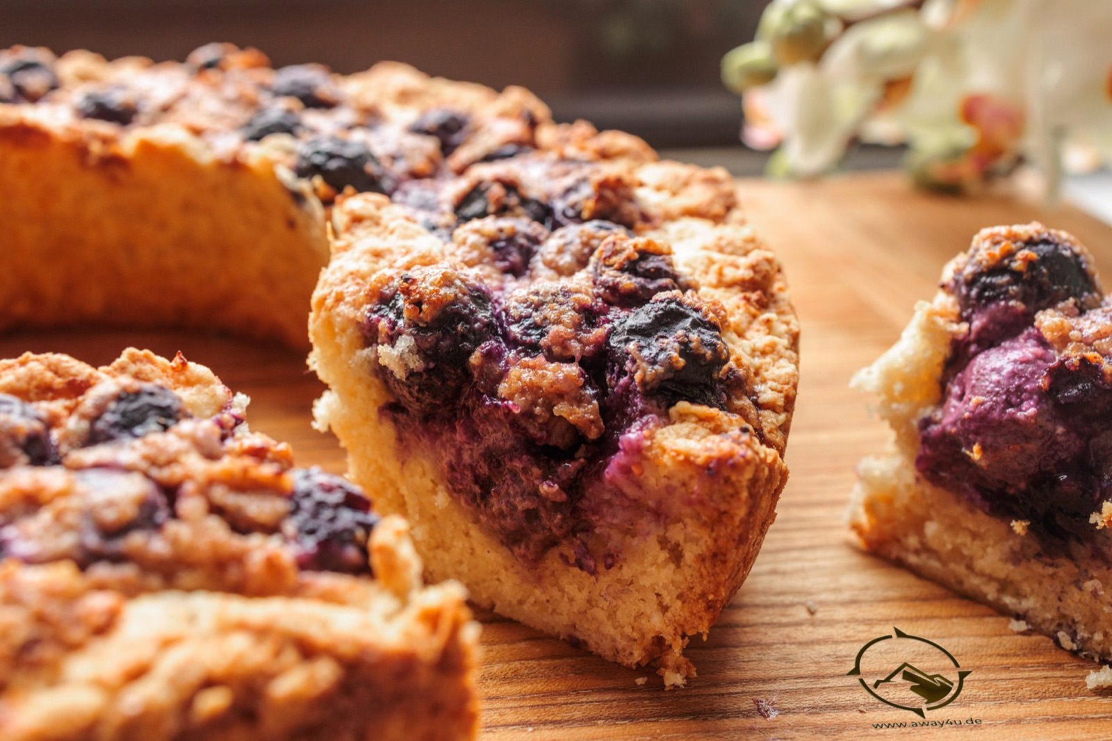 Angeschnittener Blaubeerkuchen mit Streuseln auf einem Holzbrett, am linken Rand ist die Ringform zu erkennen