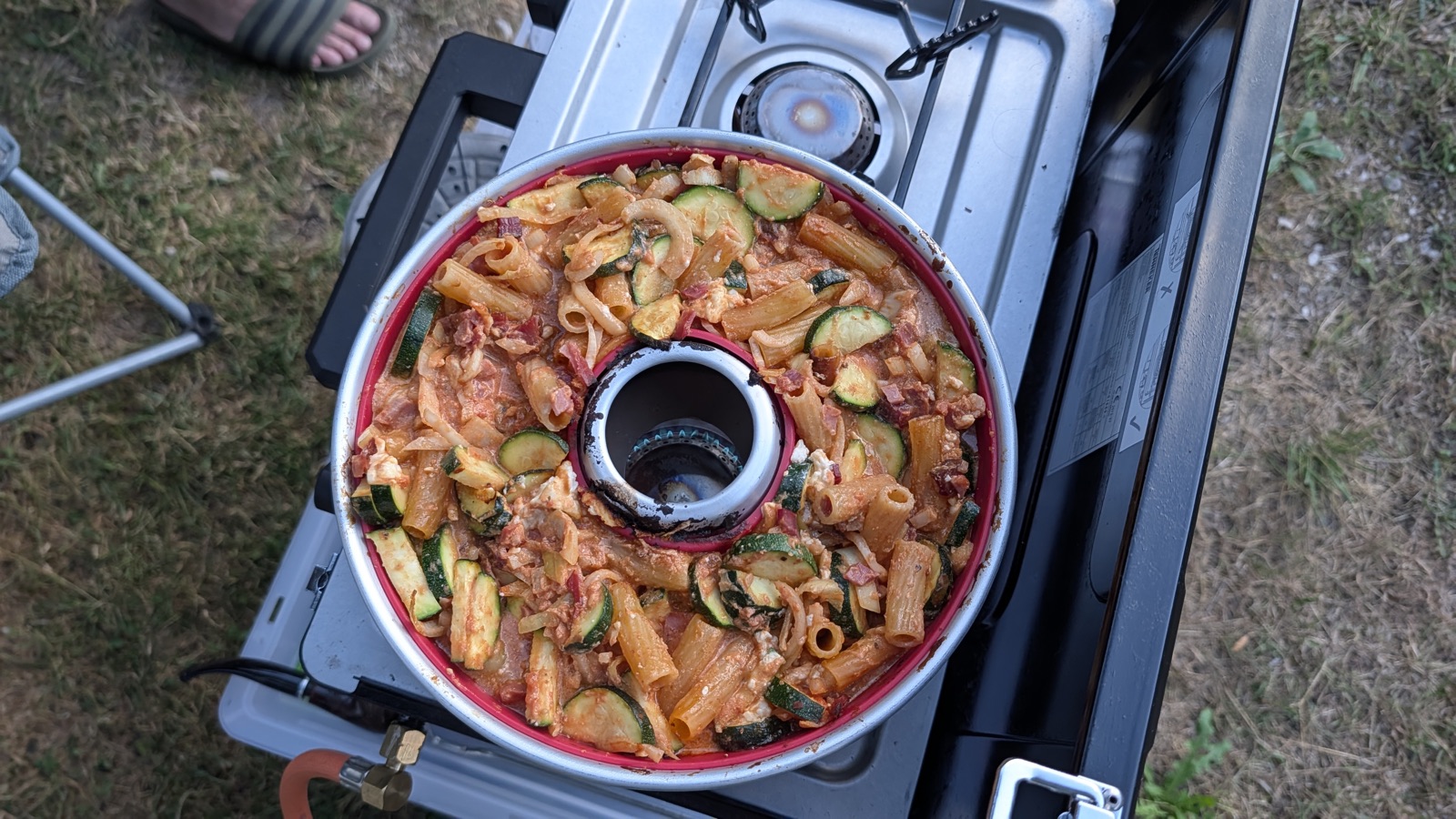 Nudelauflauf mit Zucchini und Tomatensauce in der roten Silikonform des Omnia, auf einem zweiflammigen Campinggaskocher im Gras